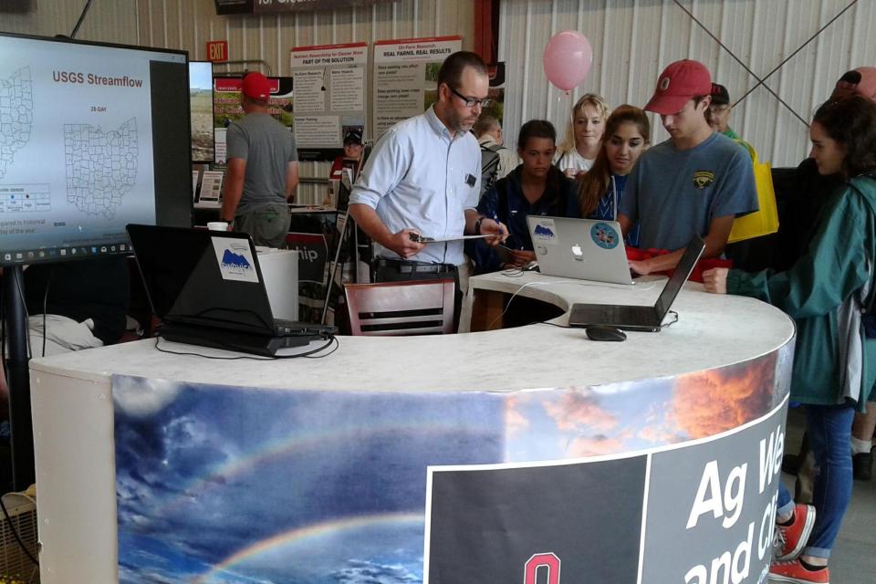 Jason Cervenec at an event with Agriculture Week. He is standing in front of a chart and working with students on a laptop.