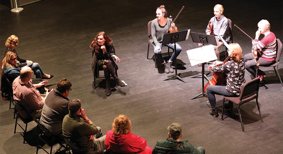 Tracie McCambridge sitting on a stage in a circle with musicians holding instruments. She is facing and talking to the crowd.
